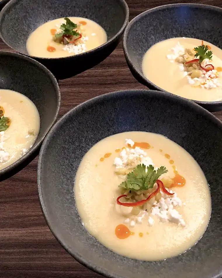 Multiple bowls of chilled corn soup plated in sequence on a walnut table