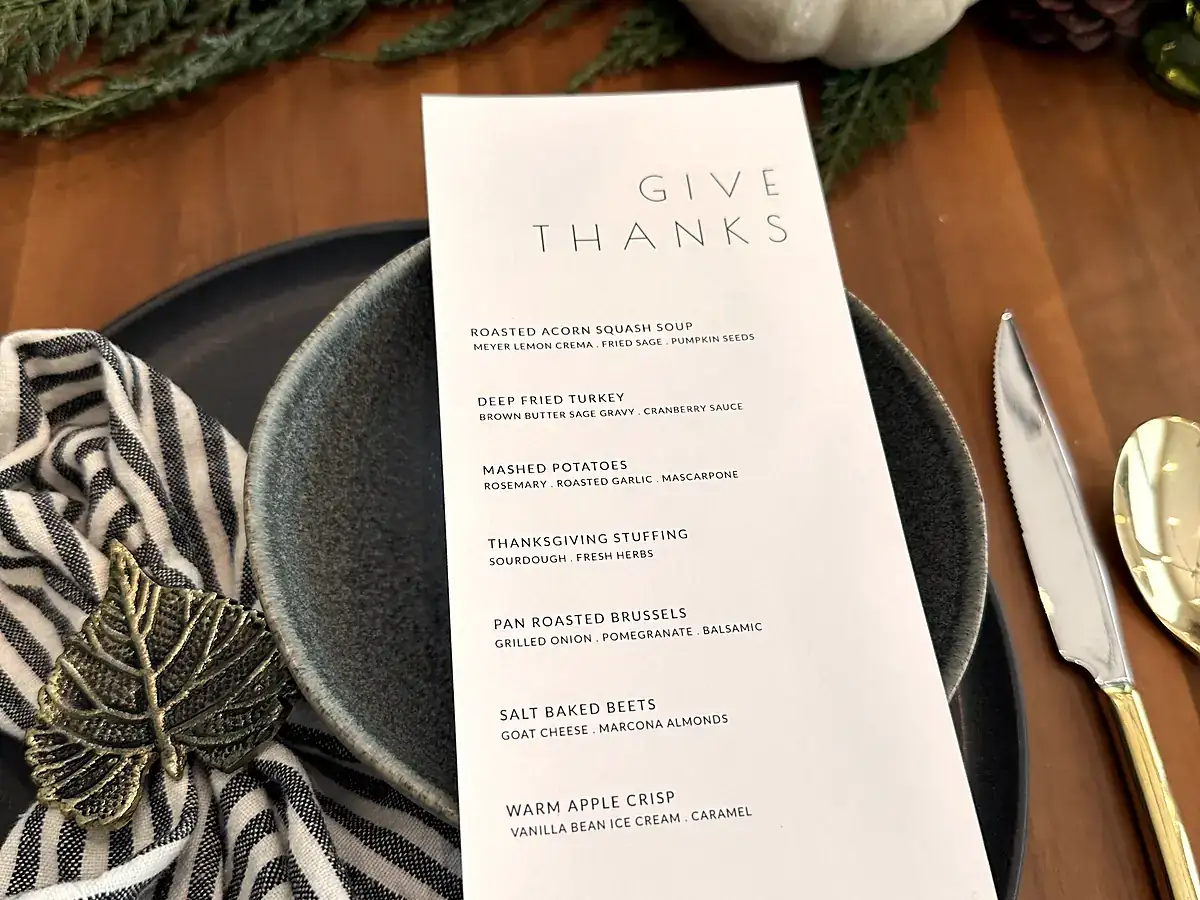 An autumn tablescape with a printed menu card, gourds, evergreen, gold flatware and a matte black charger